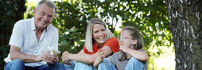 Ein älterer Mann, eine junge Frau und ein Mädchen im sitzen unter einem Baum und lachen.