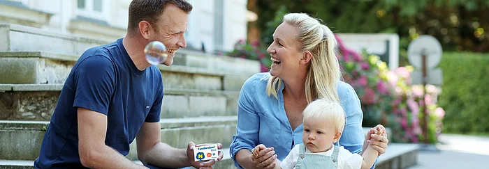 Ein Man und eine Frau sitzen im Freien auf Stufen und sehen einander an. Der Mann hält den Familienland*Pass in die Kamera. Die Frau hält ein Kleinkind.