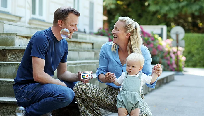 Ein Man und eine Frau sitzen im Freien auf Stufen und sehen einander an. Der Mann hält den Familienland*Pass in die Kamera. Die Frau hält ein Kleinkind.